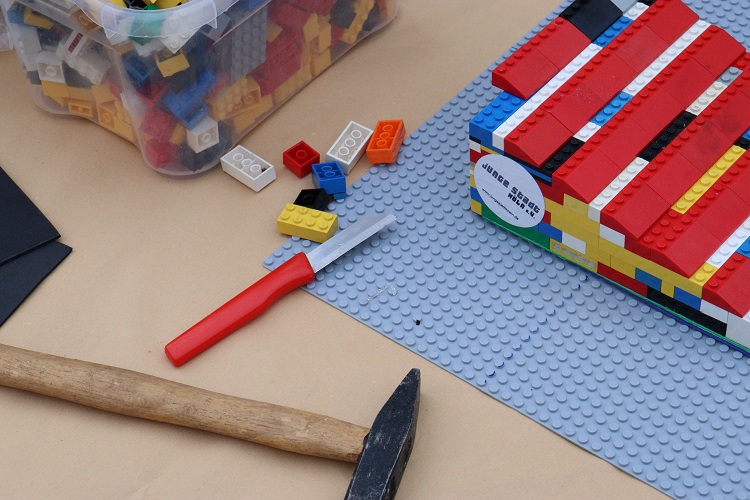 Eine Legorampenbau-Aktion mit Hammer, Cuttermesser, einzelnen Legosteinen, einer grauen Lego-Grundplatte und einer fertigen Legorampe.